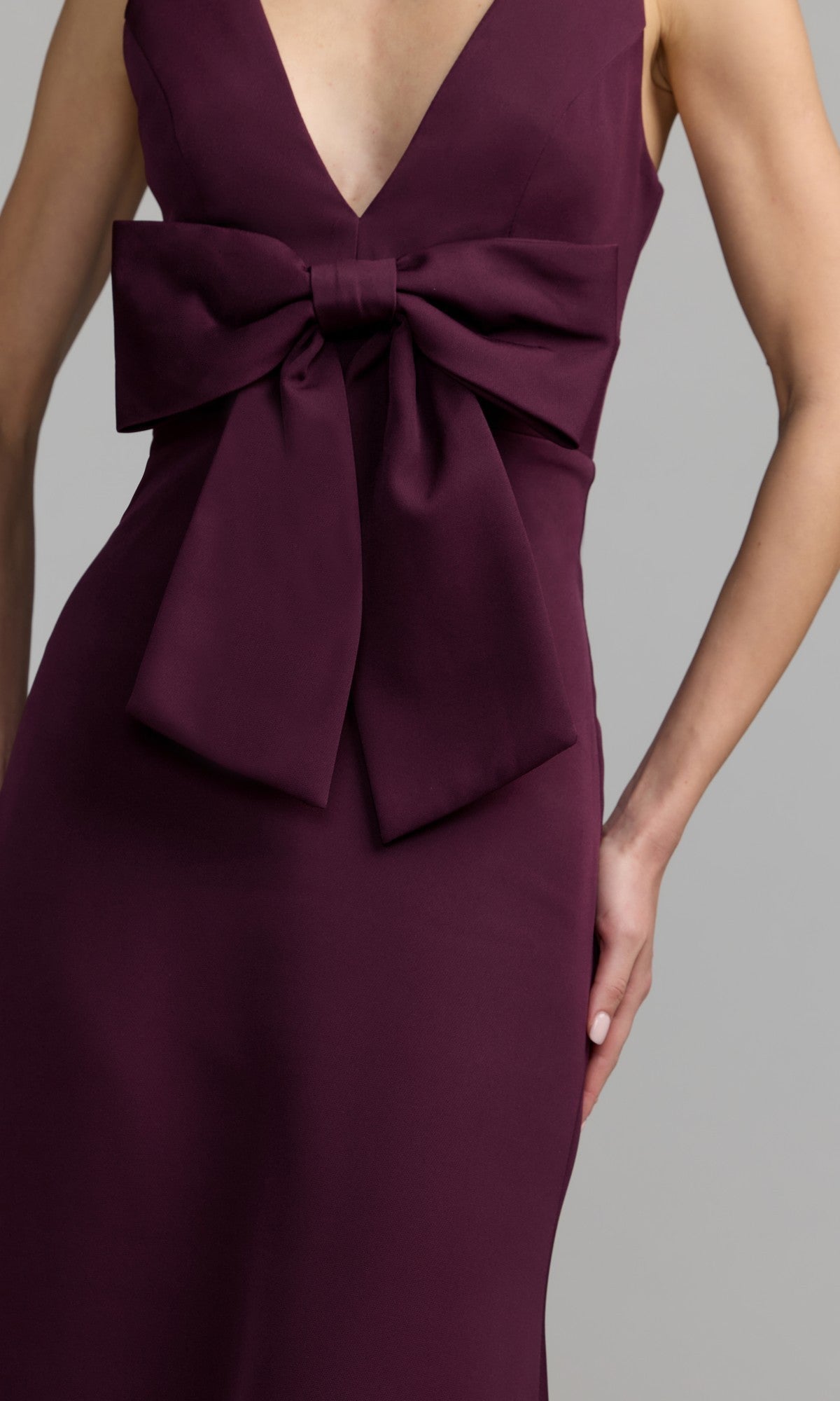 A person wearing the Mac Duggal 50087 Bow-Front Green Formal Dress, featuring a sleeveless design, deep V-neckline, and a large bow accent at the waist.